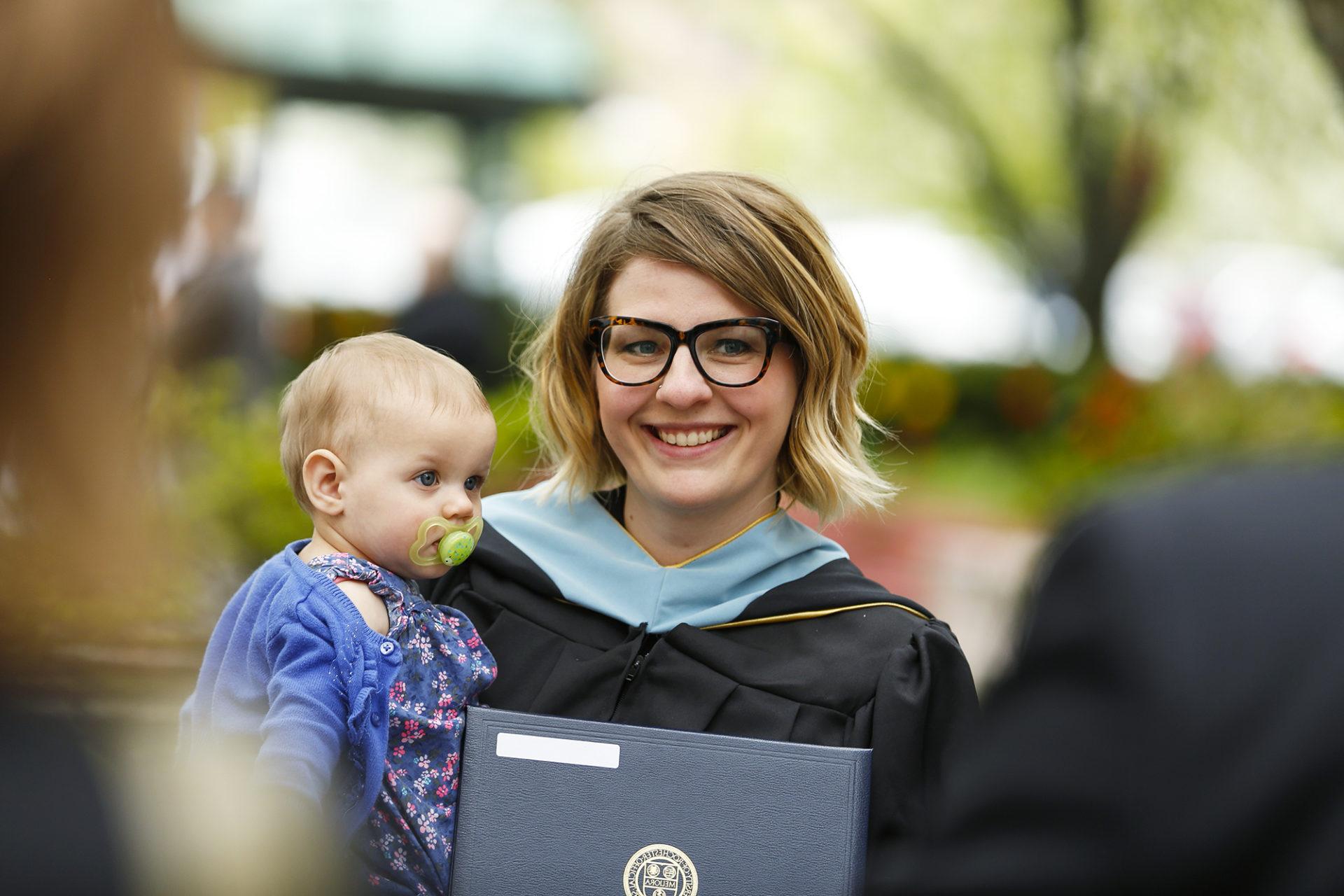 A Warner School of Education graduate poses for a photo holding her diploma and her child